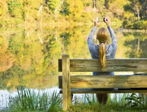 Woman sitting on bench in park in front of water and taking a picture on her phone.