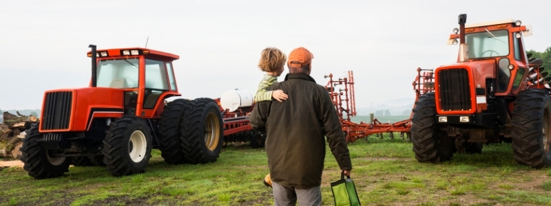 Man holding son while in field with tractors working